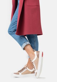 Baskets blanches et beige avec des accents marron, présentant une semelle texturée et un design à lacets, portées avec un jean bleu retroussé et un manteau rouge.