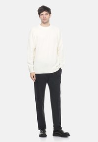 Maglione a maglia color crema con vestibilità rilassata, texture a coste e maniche lunghe, abbinato a pantaloni scuri e scarpe nere.