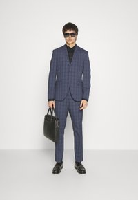 Navy checkered suit with a tailored fit, paired with a black shirt. Model holds a black leather bag and wears dark sunglasses. Black shoes.