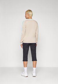 Béžový rebrovaný sveter s modrými pásikmi, doplnený o čierne skrátené nohavice. Model nosí biele tenisky a ponožky. Celkový vzhľad má hladkú textúru.