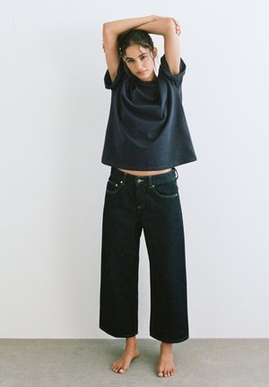 Fiatal nő mezítláb áll fehér fal mellett, laza fekete pólót és széles szárú, rövid fekete farmert visel, karjai a feje fölött keresztezetten emelve.