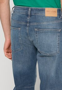 Jeans de denim azul con un lavado ligero, que cuentan con dos bolsillos traseros, un parche de marca de cuero y un diseño ajustado. Textura suave y costuras sutiles.