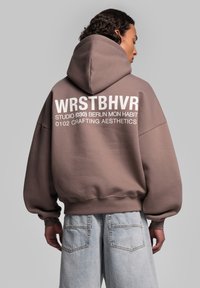 Brauner, übergroßer Hoodie mit Kapuze, der auf der Rückseite mit weißem Text bedruckt ist. Leichter Stoff und gerippte Bündchen. Kombiniert mit hellblauen Denim-Shorts.