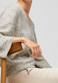V-neck pullover in light gray knit with a textured pattern. Loose sleeves and a relaxed fit. Model seated with one hand on a wooden chair.