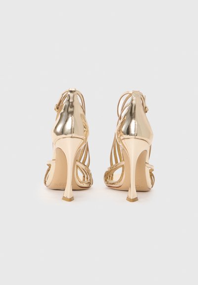 Gold high-heeled sandals with sleek metallic finish, open back, crisscross straps, and slender stiletto heels.