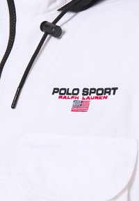 Bílá bunda s černým zipem a nastavitelnou šňůrkou. Loga "POLO SPORT" a "RALPH LAUREN" v černé a červené barvě, s designem americké vlajky.