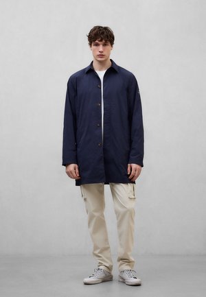 Ung mann iført en marineblå knappet frakk, hvit T-skjorte, beige cargobukser og hvite joggesko, stående mot en ensfarget grå bakgrunn.