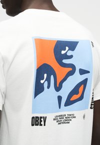 Άτομο που φοράει ένα λευκό T-shirt με αφηρημένο μπλε και πορτοκαλί γραφικό και τη λέξη "OBEY" μαζί με μεγάλα ονόματα πόλεων στην πλάτη.