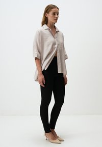 Camicia di lino beige chiaro con colletto, apertura frontale a bottoni e maniche arrotolate, abbinata a leggings neri e tacchi a punta nudi.