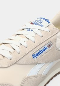 Bēšas zamšādas kedas ar baltām auklām, Reebok logo un zilu akcentu. Iespējojums ir gluda ādas strīpa un teksturēta gumijas zole.