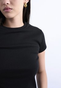 Γυναίκα που φοράει μια απλή μαύρη μπλούζα και στριφογυριστά χρυσά κρίκους, εμφανιζόμενη από τον λαιμό έως το μέσο του κορμού μπροστά σε λευκό φόντο.