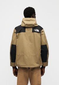 Αδιάβροχο μπουφάν με μπεζ σώμα, μαύρα επάνω πάνελ και κουκούλα. Διακριτικό λογότυπο στην πλάτη και ραφές με αντίθετο χρώμα.