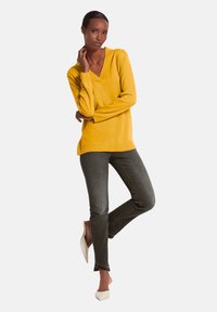 Gelbes V-Ausschnitt-Pullover mit langen Ärmeln, kombiniert mit dunkelgrauen Jeans und beigen Absätzen. Stricktextur, lockere Passform und klare Linien.