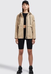 Beige kapucnis dzseki, két elülső zsebbel, fekete fűzőkkel és textúrált ujjal, fekete crop top és sort felett viselve. Fekete cipő.