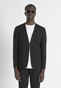 Blazer nero su misura con scollatura a V, due bottoni e tasche frontali. Indossato sopra una t-shirt bianca, abbinato a pantaloni neri e occhiali da sole.