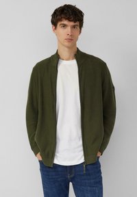 Olijfgroene cardigan met ritssluiting en geribbelde accenten, gemaakt van een zachte gebreide stof; gedragen over een wit T-shirt en een blauwe spijkerbroek.