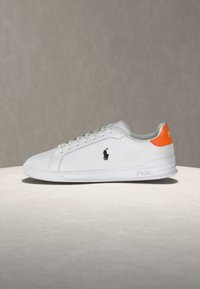 Λευκό δερμάτινο αθλητικό παπούτσι με κορδόνια, μαύρο λογότυπο πόλο στο πλάι και πορτοκαλί ταινία στη φτέρνα, τοποθετημένο σε ουδέτερη επιφάνεια.
