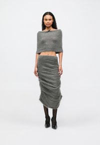 Femeie îmbrăcată cu un top scurt off-shoulder, căptușit de un gri închis, și o fustă midi cu talie înaltă asortată, ciorapi negri și tocuri negre, stând pe fundal alb.