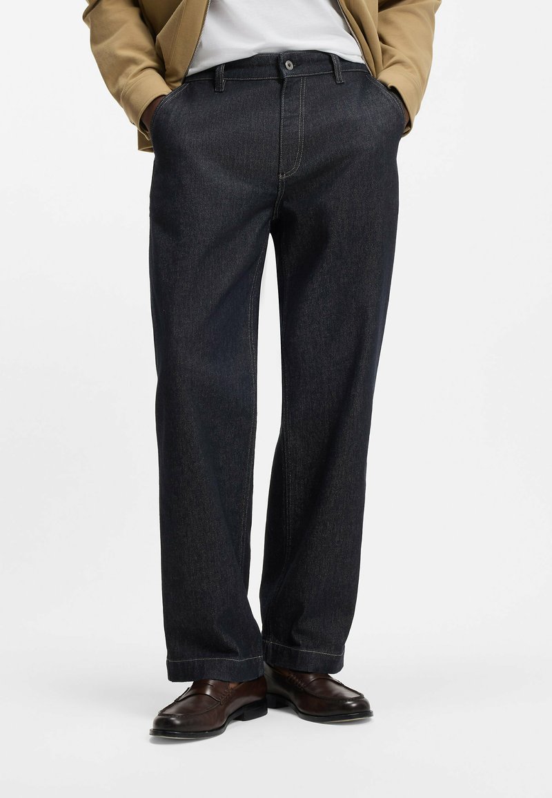 Pantaloni in denim blu scuro con taglio dritto, caratterizzati da cuciture visibili, passanti per cintura e tasche laterali. Abbinati a scarpe eleganti marroni.