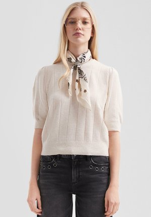 Pull court en crème clair à manches courtes avec texture côtelée, encolure ronde et taille cintrée. Associé à un foulard à motifs et un jean noir.