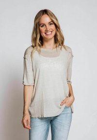 Helle beigefarbene, locker sitzende Bluse mit Ellenbogen-langen Ärmeln, rundem Ausschnitt, subtiler Textur und rohen Kanten, kombiniert mit hellblauen Jeans.