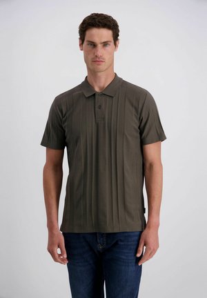 Mann mit lockigem dunklem Haar, der ein dunkelolivfarbenes kurzärmeliges Poloshirt und blaue Jeans vor einem schlichten hellen Hintergrund trägt.
