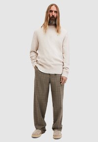 Hellbeige gestrickter Pullover, lockere Passform, kombiniert mit braunen Karohosen. Model trägt beiges Schuhwerk. Grauer Hintergrund.