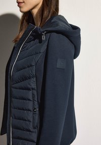 Giacca ibrida blu navy con parte frontale imbottita e cappuccio, caratterizzata da un motivo trapuntato texturizzato e lati in maglia liscia, accentuata da un'etichetta con logo.