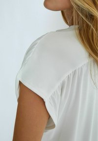 Blusa bianca con maniche corte leggermente arricciate. Tessuto liscio con una vestibilità comoda e scollo rotondo, che mette in mostra un materiale morbido e leggero.