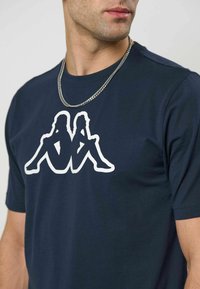 Mann iført marineblå t-skjorte med hvitt logo av to sittende figurer rygg mot rygg og et sølvkjede rundt halsen