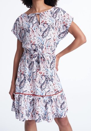 Femme portant une robe blanche à manches courtes avec des motifs cachemire bleus et rouges, une ceinture à la taille et un ourlet froncé.
