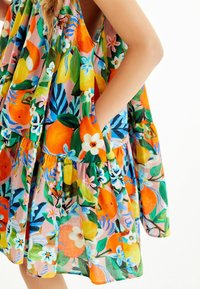 Blumenkleid mit lebendigen orange- und gelbfarbigen Obstmustern, grünen Blättern und einem hellrosa Hintergrund. Mit Taschen und geschichteten Rüschen.