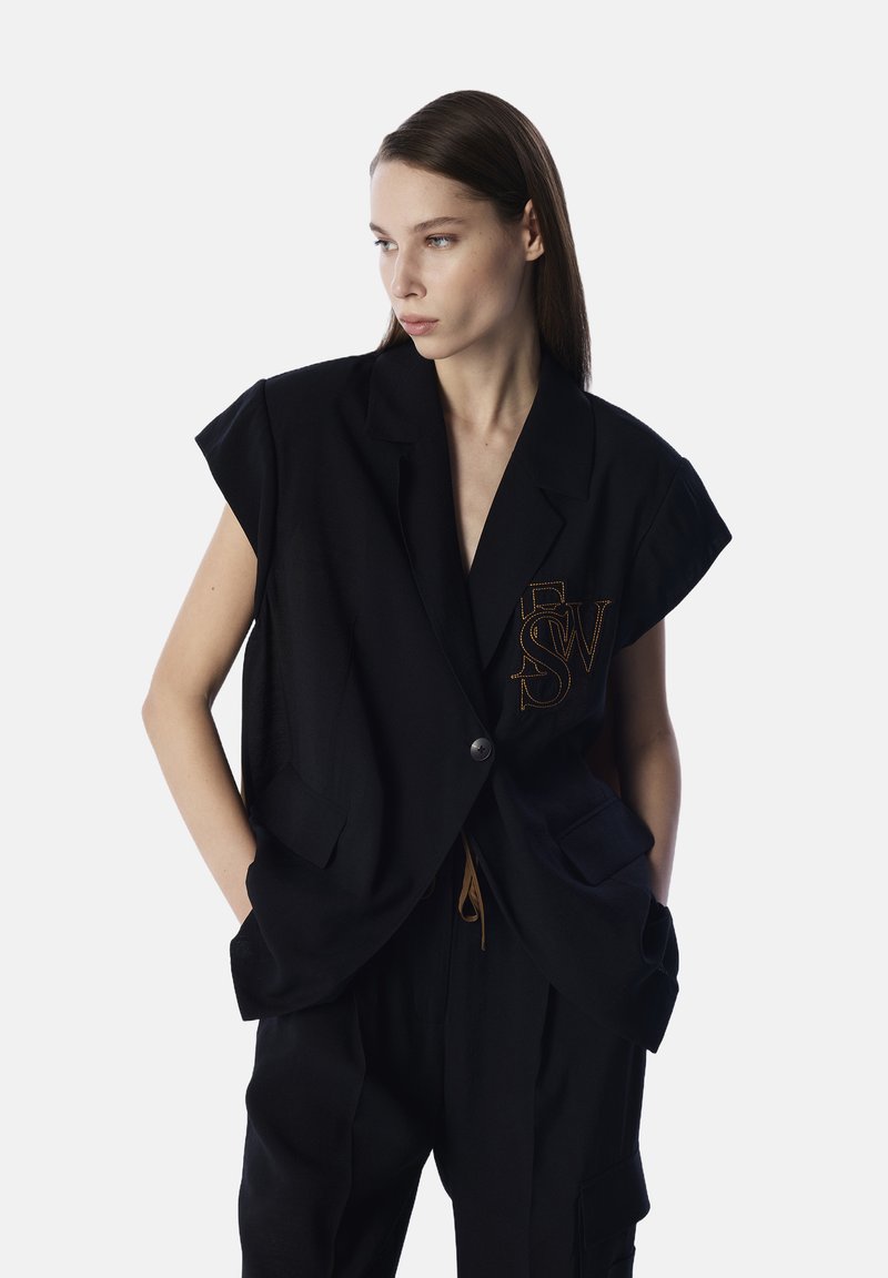 Czarna, oversize kamizelka z krótkimi rękawami, z przodu zapinana na guziki i z bocznymi kieszeniami. Zawiera wyhaftowane inicjały w kolorze pomarańczowym.