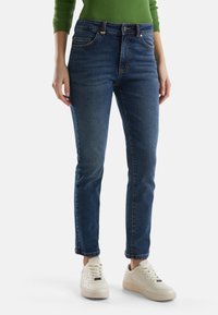 Donkerblauwe denimspijkerbroek met een rechte pasvorm, vijfzakkenontwerp, zichtbare stiksels en opgerolde boorden. Gecombineerd met witte sneakers.