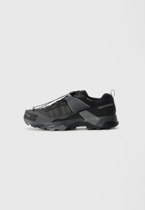 Chaussure d'extérieur noire avec un dessus texturé, système de laçage élastique, coque de protection renforcée à l'avant, et semelle extérieure robuste pour une bonne traction. Présente un détail de logo.