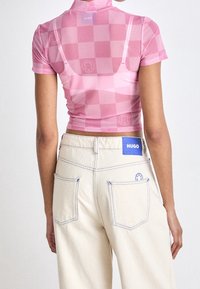 Růžový kostkovaný cropped top s vysokým límcem; spárovaný s béžovými džíny s modrým prošíváním a štítkem loga na zadní kapse.