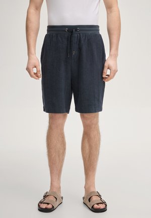 Mężczyzna ubrany w granatowe szorty ze ściągaczem i beżowe sandały z klamrą, stojący na tle jednolitego jasnego tła.