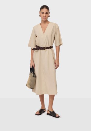 Kobieta ubrana w beżową sukienkę midi z dekoltem w serek i szerokimi rękawami, z ciemnym plecionym paskiem, brązowymi sandałami, kolczykami kołami oraz trzymająca paskowany pleciony torebkę.