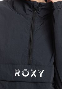 Svart vindjacka med hög krage, halvdragkedja och en tydlig vit "ROXY"-logotyp över bröstet. Slät nylontextur.