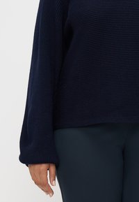 Pull en maille bleu marine avec des manches amples et une texture côtelée, associé à un pantalon ajusté vert foncé.