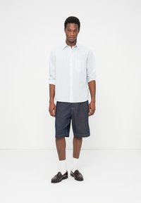 Άνδρας που στέκεται μπροστά σε λευκό φόντο, φορώντας ανοιχτό μπλε πουκάμισο με μακριά μανίκια, σκούρες τζιν βερμούδες, λευκές κάλτσες και σκούρα καφέ μοκασίνια.