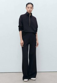 Sweater en polaire noir à col zippé avec une coupe décontractée, associé à un pantalon noir à jambes larges. Texture lisse et design minimaliste, rehaussé par des chaussures simples.