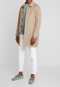 Abrigo a cuadros beige con cierre de botones, combinado con un suéter azul claro, pantalones blancos y mocasines de ante verde. Textura suave, corte estructurado.