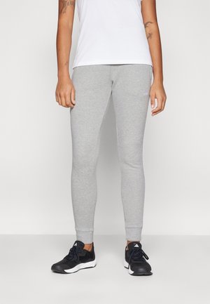 Personne portant un pantalon de jogging slim gris clair, une chemise blanche et des baskets noires avec des semelles blanches, debout devant un fond blanc uni.