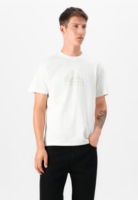 T-shirt branca de algodão com um logo Quiksilver verde claro, gola redonda, mangas curtas, ajuste solto, usada com calças pretas, textura mínima.