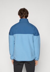 Giacca in pile con colletto alto, presenta la parte inferiore di colore blu chiaro e la parte superiore di colore blu scuro, tessuto testurizzato e dettagli di cucitura a contrasto.