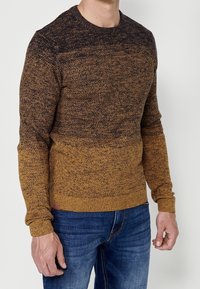 Homme portant un pull en maille dégradé marron et noir et un jean bleu, debout devant un fond clair uni.
