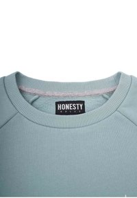 Vihreä svetari, jossa on ribbireuna kauluksessa ja hihoissa, pehmeä rakenne, ja sisäpuolella etiketti, jossa lukee "REHELLISYYS ON SÄÄNTÖ".