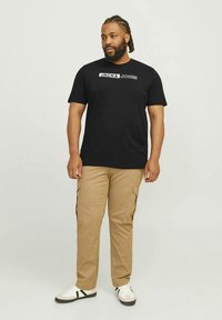 Juoda medvilninė marškinėliai su „JACK & JONES“ logotipu, priderinti prie žalvarinių kargo kelnių su šoninėmis kišenėmis ir baltais sportbačiais su juodomis detalėmis.