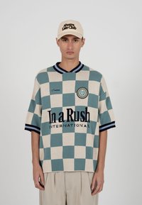 Ung mand iført beige kasket med teksten "Always Late Club" og oversized kortærmet ternet skjorte med teksten "In a Rush International".